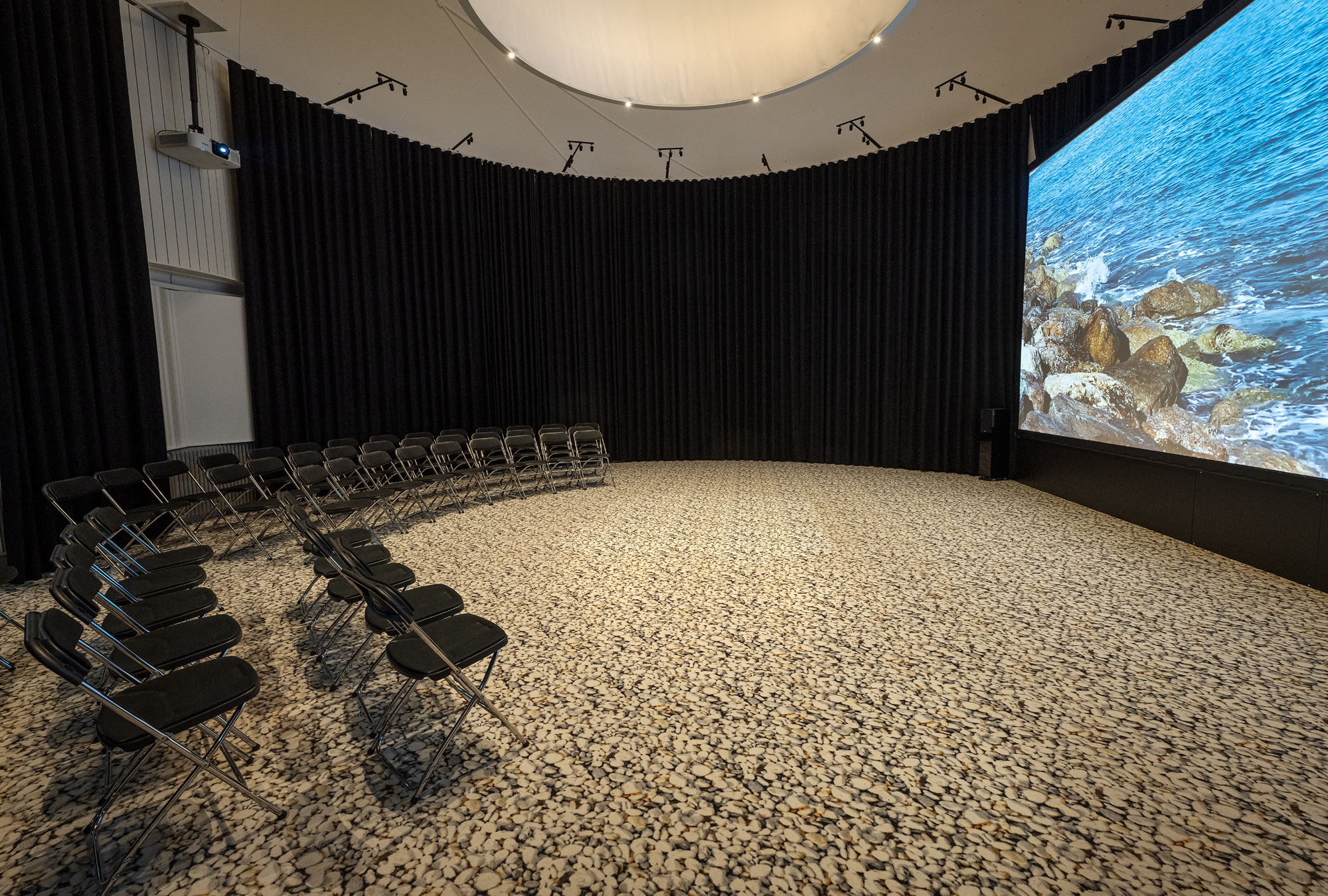 En rund sal med kupolformat tak. På högra sidan är en stor projektorskärm och till vänster många stolar i rader.