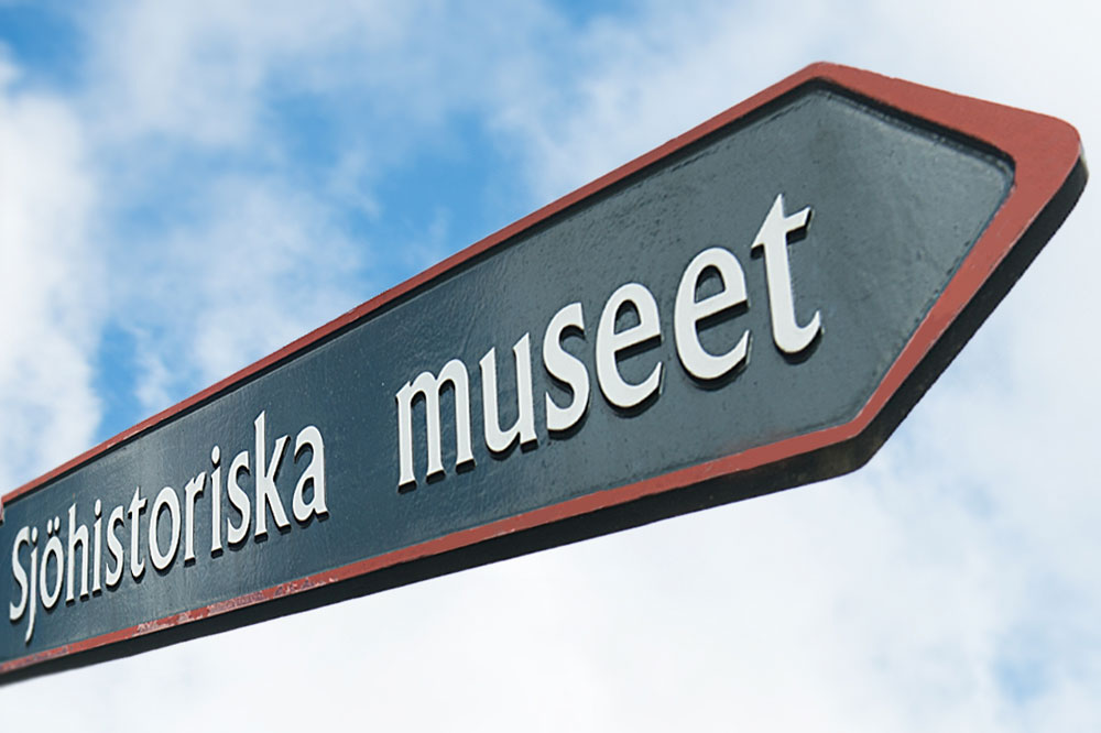 Skylt i form av en pil. På skylten står Sjöhistoriska museet. Ovanför skylten blå himmel med sommarmoln. 
