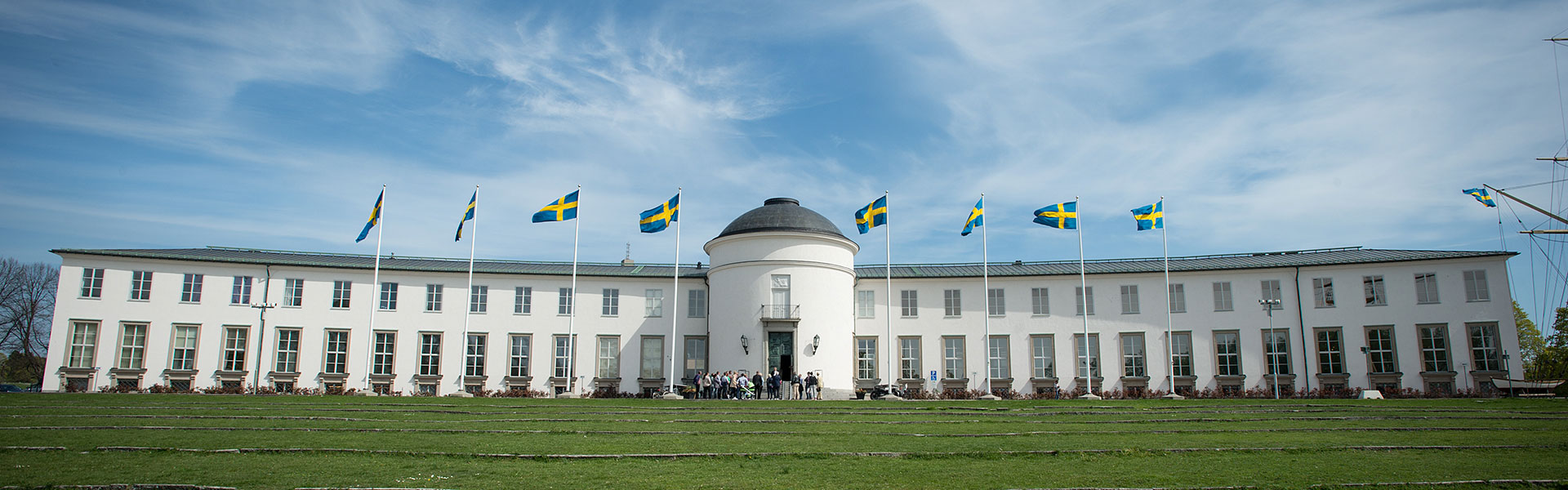 Sjöhistoriska museet framifrån. En grupp människor står nedanför museets entrétrappa. Flaggstänger framför museet med hissade svenska flaggor.
