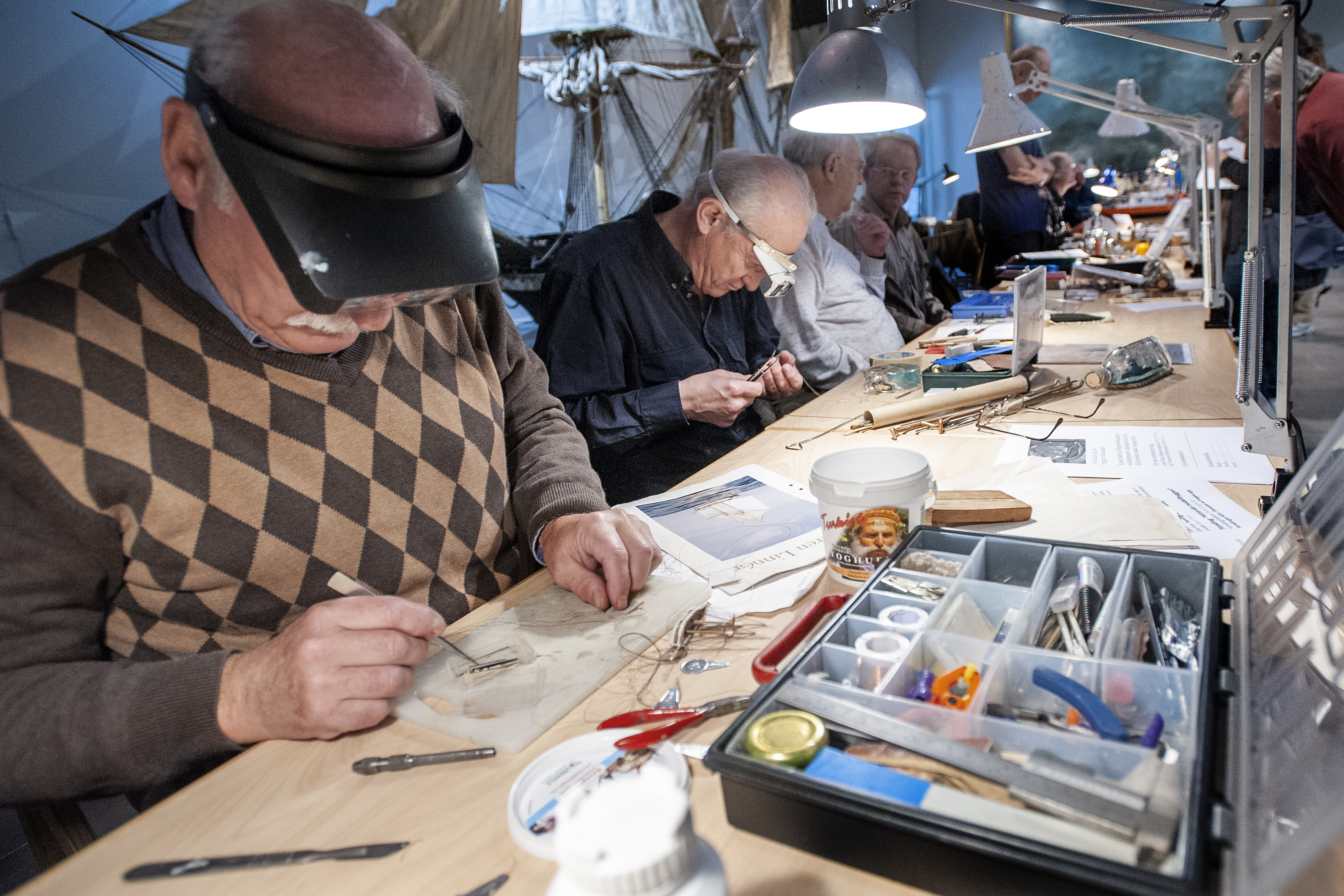 Bilden skildrar i förgrunden en modellbyggare som jobbar under stark belysning i bakgrunden flera modellbyggare i arbete som sitter längsmed varandra. ""