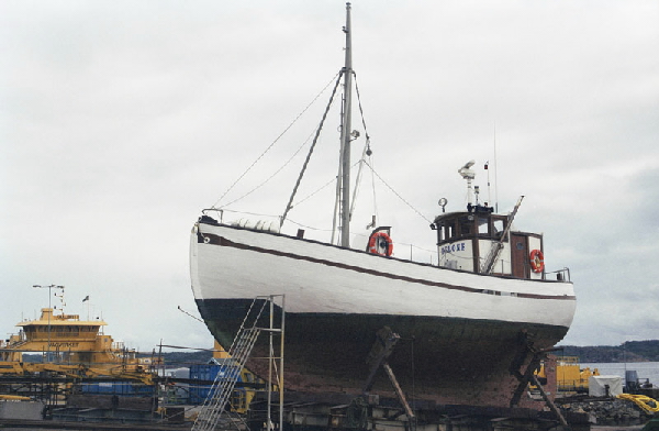 Fiskefartyget Belone ligger på torra land med det svartvita skrovet synligt. Hon har en styrhytt i aktern och en mast i fören. Hon har två livbojar för säkerhets skull.