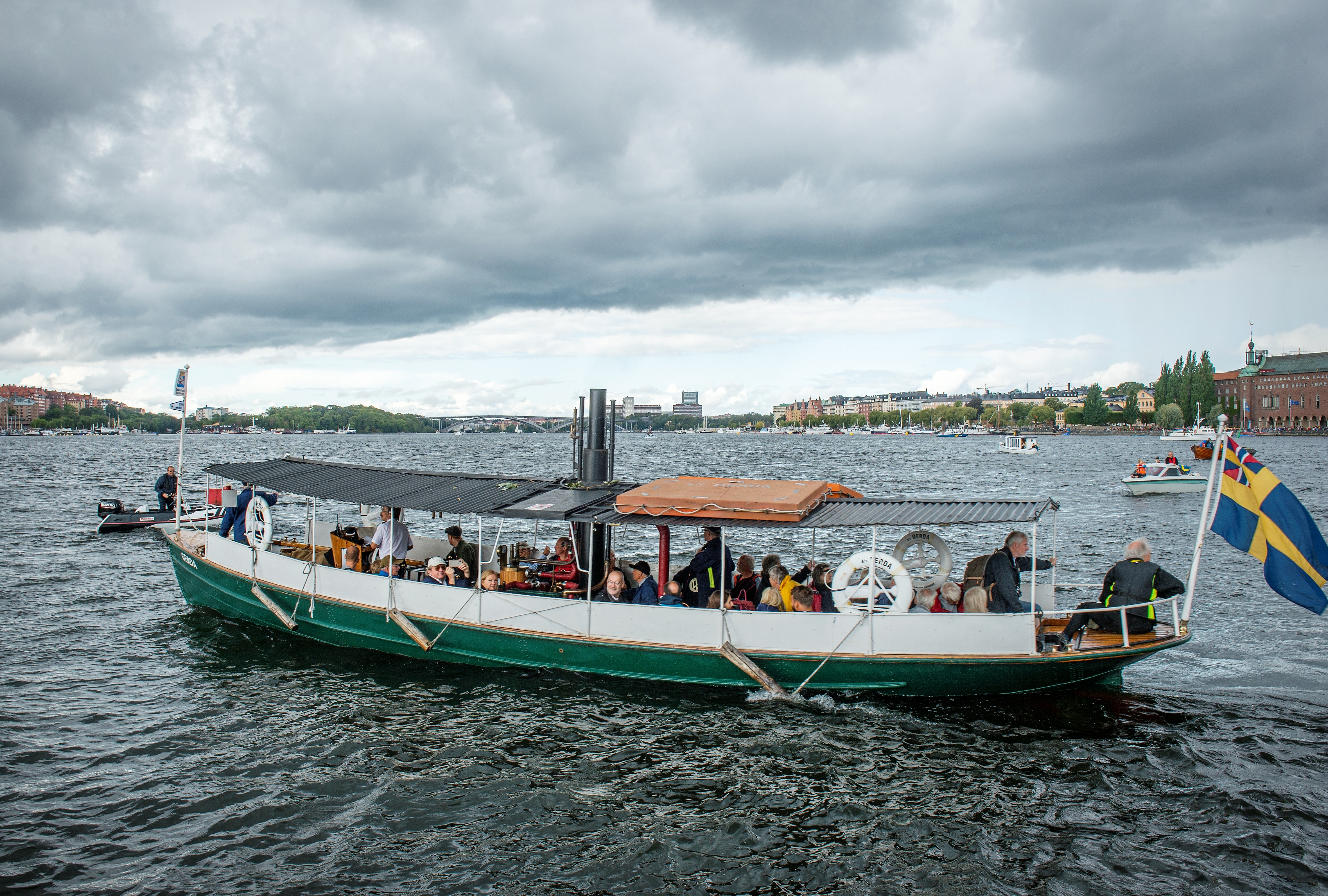 Gerda är en öppen låg ångslup med enkelt plåttak på stolpar. Skrovet är grönmålat med svart skorsten. På bilden är båten fylld med passagerare och i bakgrunden skymtar Riddarfjärden och Stockholms stadshus.  