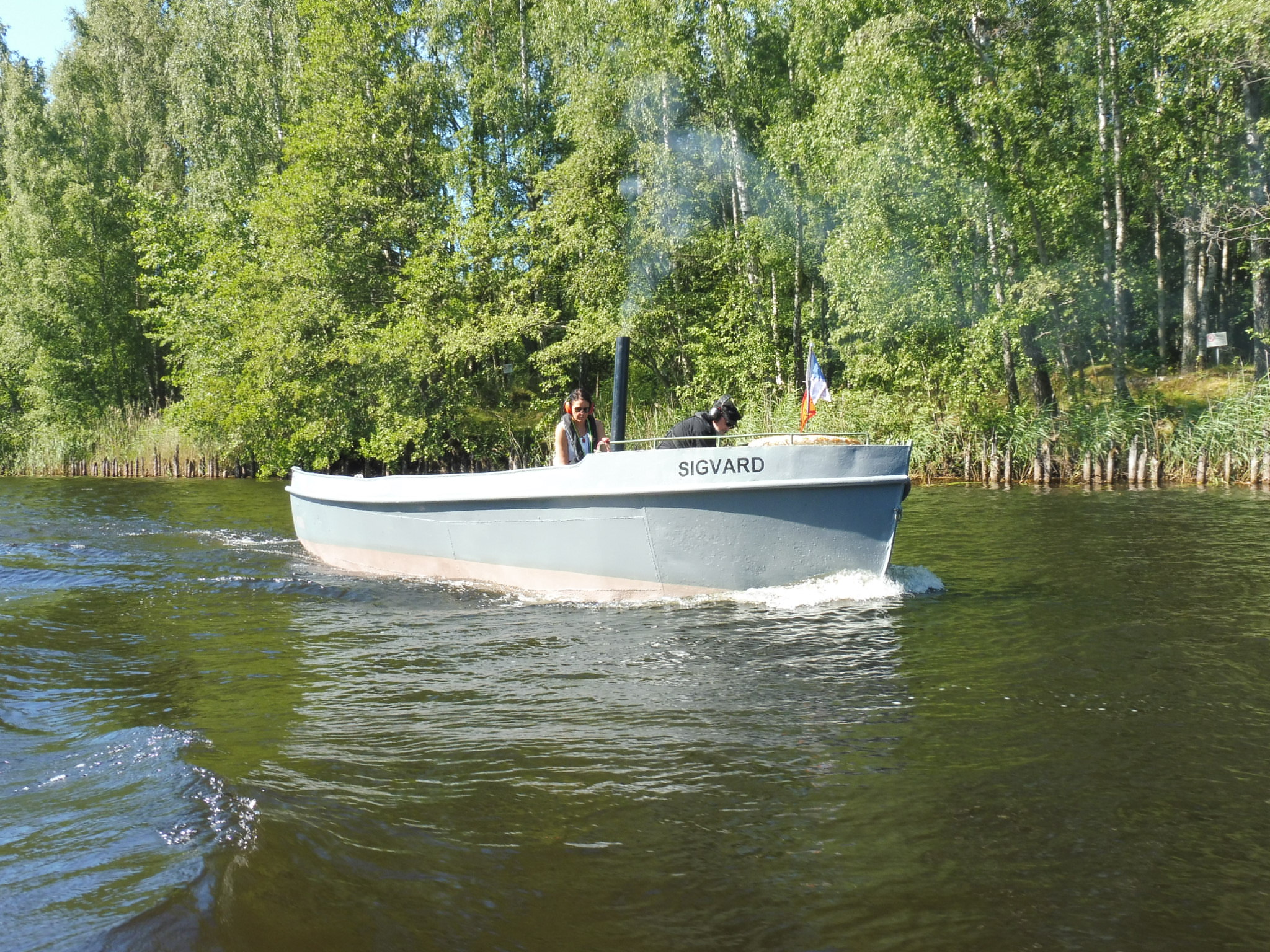 Sigvard är en liten motorvarpbåt med grått plåtskrov och en svart tunn skorsten som sticker upp midskepps. På bilden är båten ute till sjöss på en insjö. 