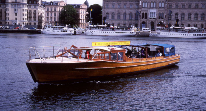 Stegeholm är en låg sightseeingbåt i ljusbrun mahogny. I fören syns ett däck med mantåg, midskepps en låg styrhytt och i akter en salong med blått kapelltak. På bilden ser man fartyget på Stockholms ström med Nationalmuseum i bakgrunden. 