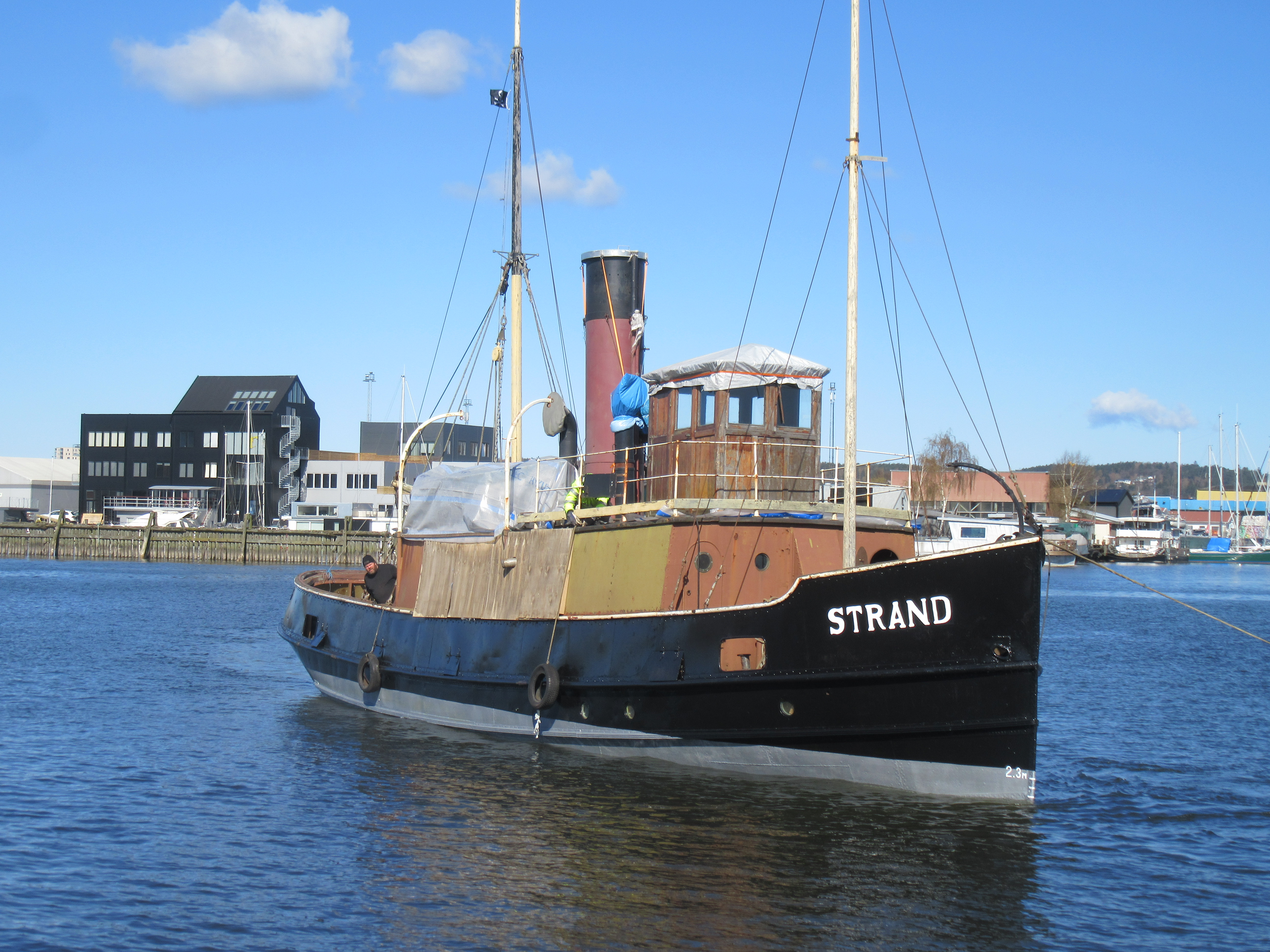 Strand är en bogserbåt med svartmålat plåtskrov och överbyggnad i olika bruna nyanser. Styrhytten är klädd i trä med en rödsvart skorsten bakom. Båten har två master.