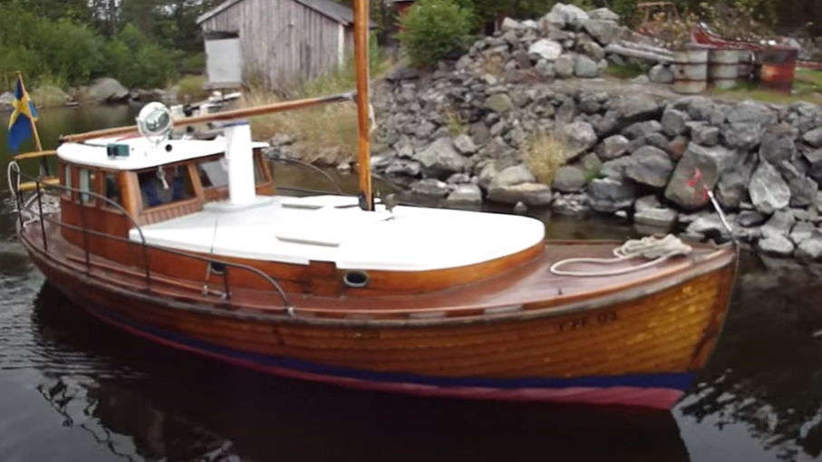 Ymer är en lotsbåt med ljusbrunt träskrov. Taket på styrhytt och ruff är vitmålat. Båten har mast och bom i trä. På bilden är fartyget på väg ut ur en naturhamn i Västernorrland. 