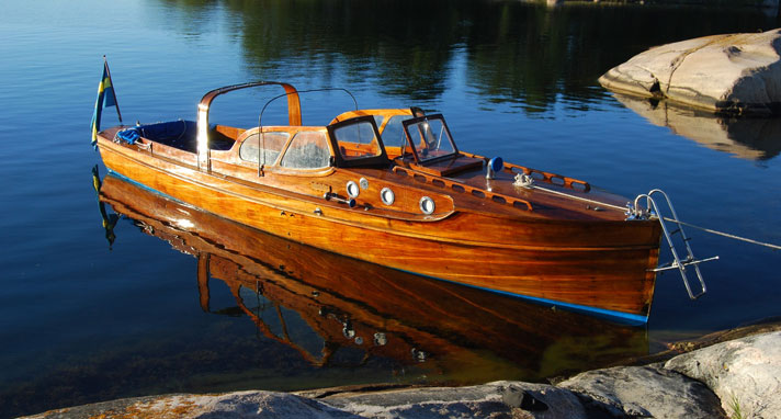 Agnes II är en ruffad motorbåt i mahogny med sittbrunn i aktern. En akterbåge för att stadga kapellet syns på bild. Båten ligger för ankar vid några klippor i spegelblankt hav.