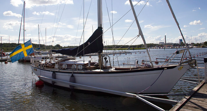 Beguine är en segelbåt som ligger förtöjd vid ett par klippor på spegelblankt hav. Dess vita skrov och de röda detaljerna skiner i solljuset. Överbyggnaden är av teak och ruffen har flertalet ventiler längs med sidorna. 