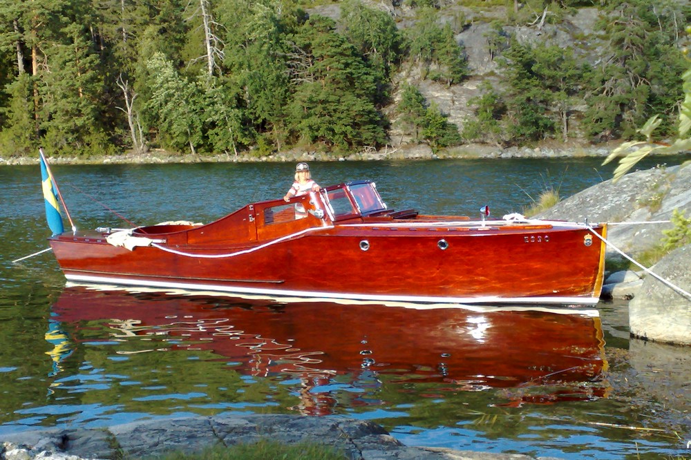 Esso är en motorbåt med rödbrun hondurasmahogny som glänser som en ädelsten! Hon har en förruff och sittbrunn med styrplats och vindruta. En person står vid styrplatsen.