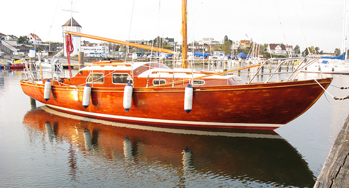 Malou är en segelbåt i mahogny med vita detaljer. Det finns inga segel i riggen och hon ligger förtöjd vid en brygga.