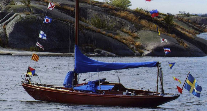 Trio är en kustkryssare i mahogny med blåa kapell. Seglet är revat men flaggspelet spelar i vinden. Hon ligger förtöjd i skärgården.