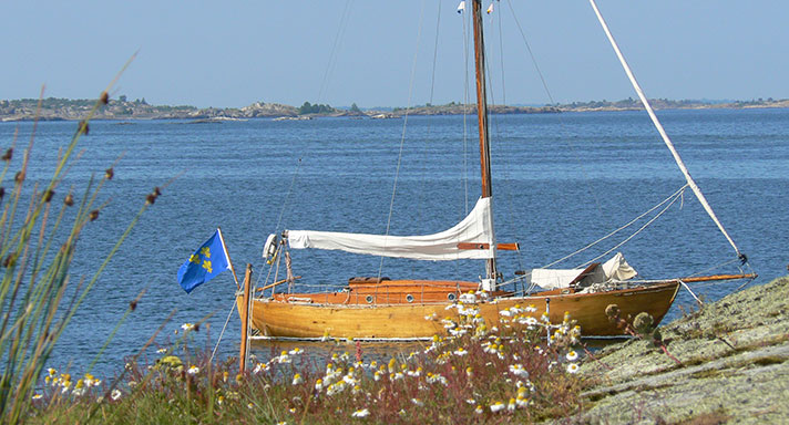 Varuna är en havskryssare med skrov i ek och överbyggnad i mahogny. Hon har seglen i sina vita kapell och ligger för ankare vid några klippor.
