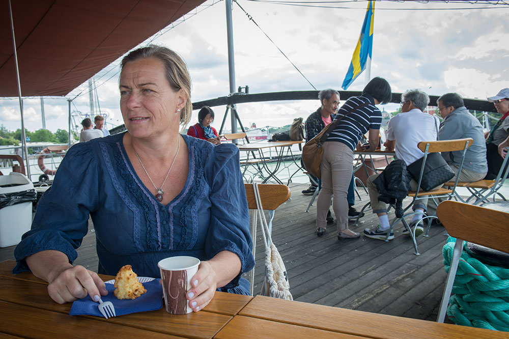 Kinna sitter vid ett cafébord ombord på Sankt Eriks aktredäck. Framför sig har hon en kopp kaffe och ett fat med en kokostopp. Bakom andra personer vid kafébord.