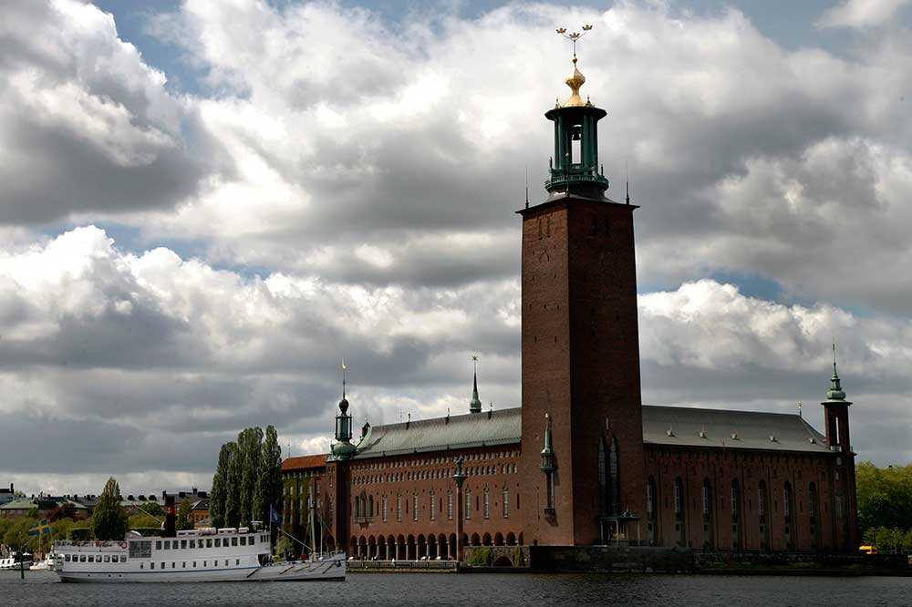 Stockholms stadshus röda tegelbyggnad vid vattnet. Överst på tornet sitter de tre gyllene kronorna. Framför passerar en vit båt.