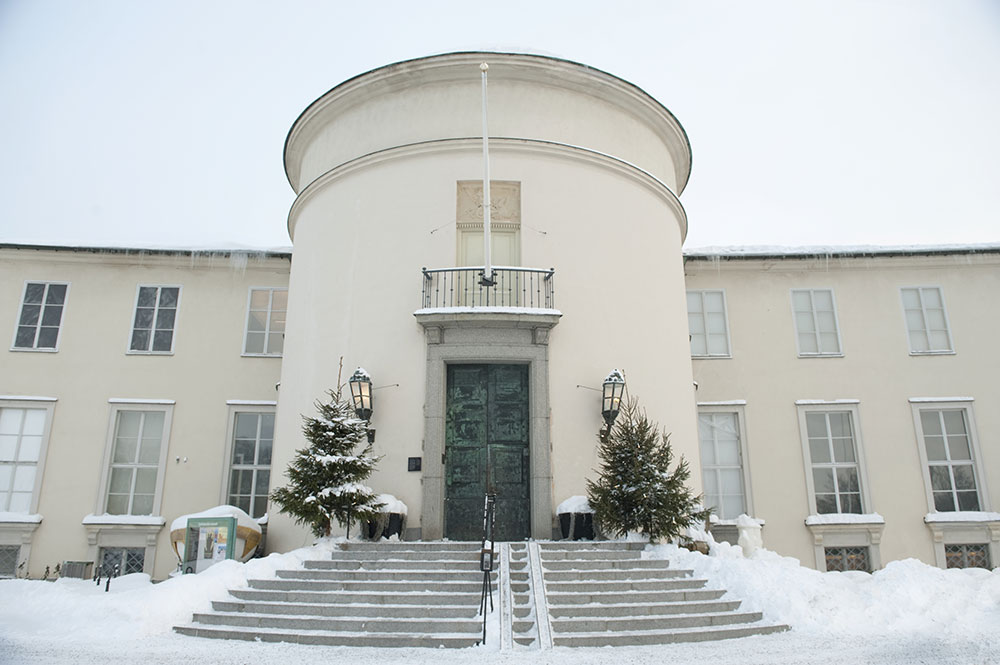 Bild på Sjöhistoriska museets entré vintertid. Sjöhistoriska museet vintertid. På marken och trappan upp till entrédörren ligger snö. Istappar hänger från taket. Två julgranar står på var sida om entrédörren.